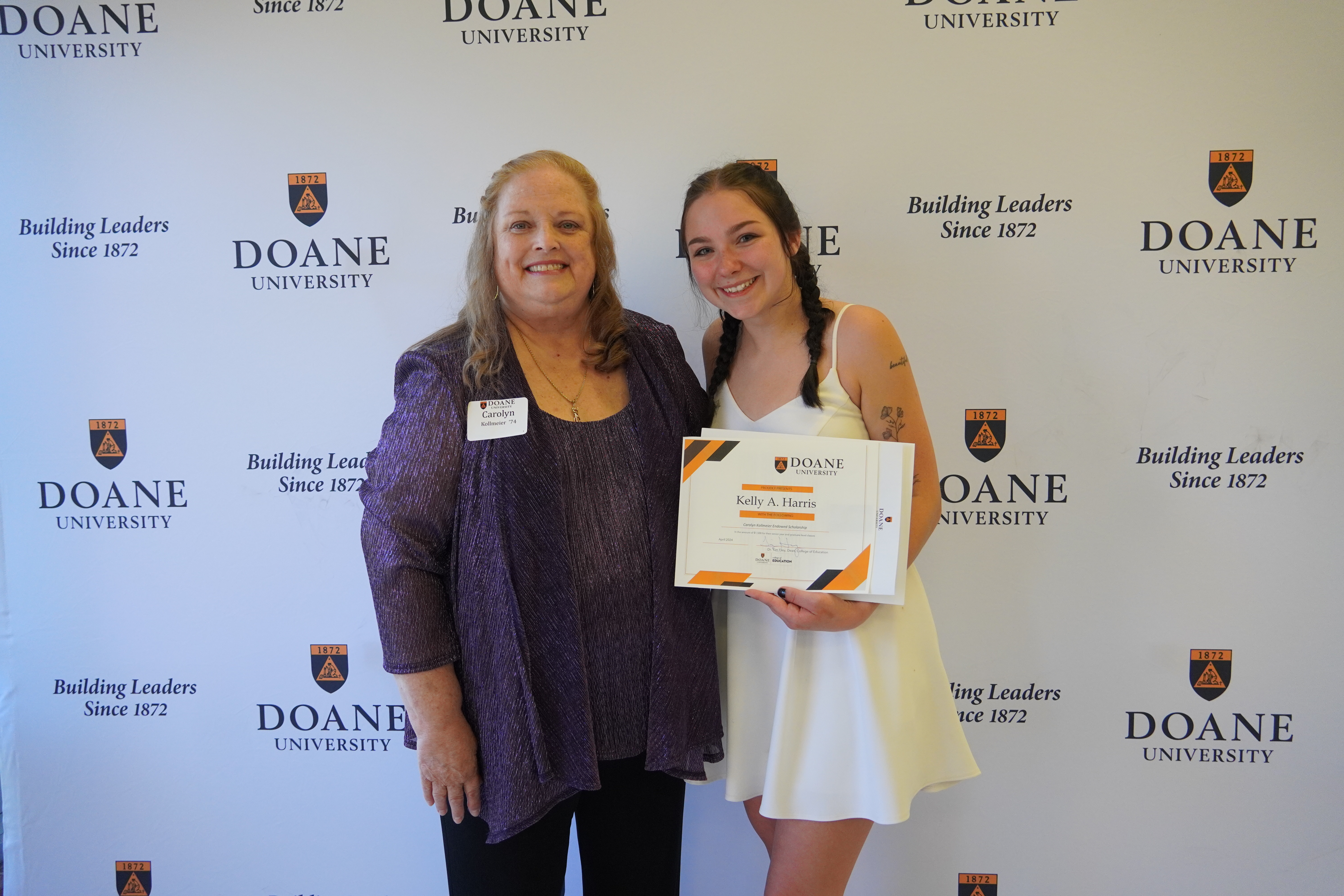 Two women, one in a purple shirt and jacket, and one in a short white dress, stand in front of a white background with text that reads "Doane University, Building Leaders Since 1872." 