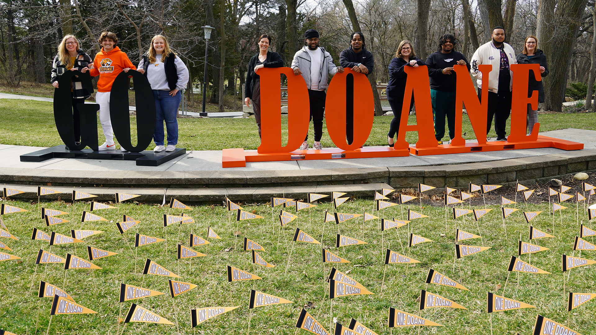 10 people, five Doane staff members, and five students, pose in front of large letters that read G-O D-O-A-N-E. The letters are black and orange and are raised on the stage of Cassel Outdoor Theatre. In front of the stage are many orange pennants on sticks that have been placed in the green grass and read “One Doane,” a nod to the name of the event — One Day. One Doane.