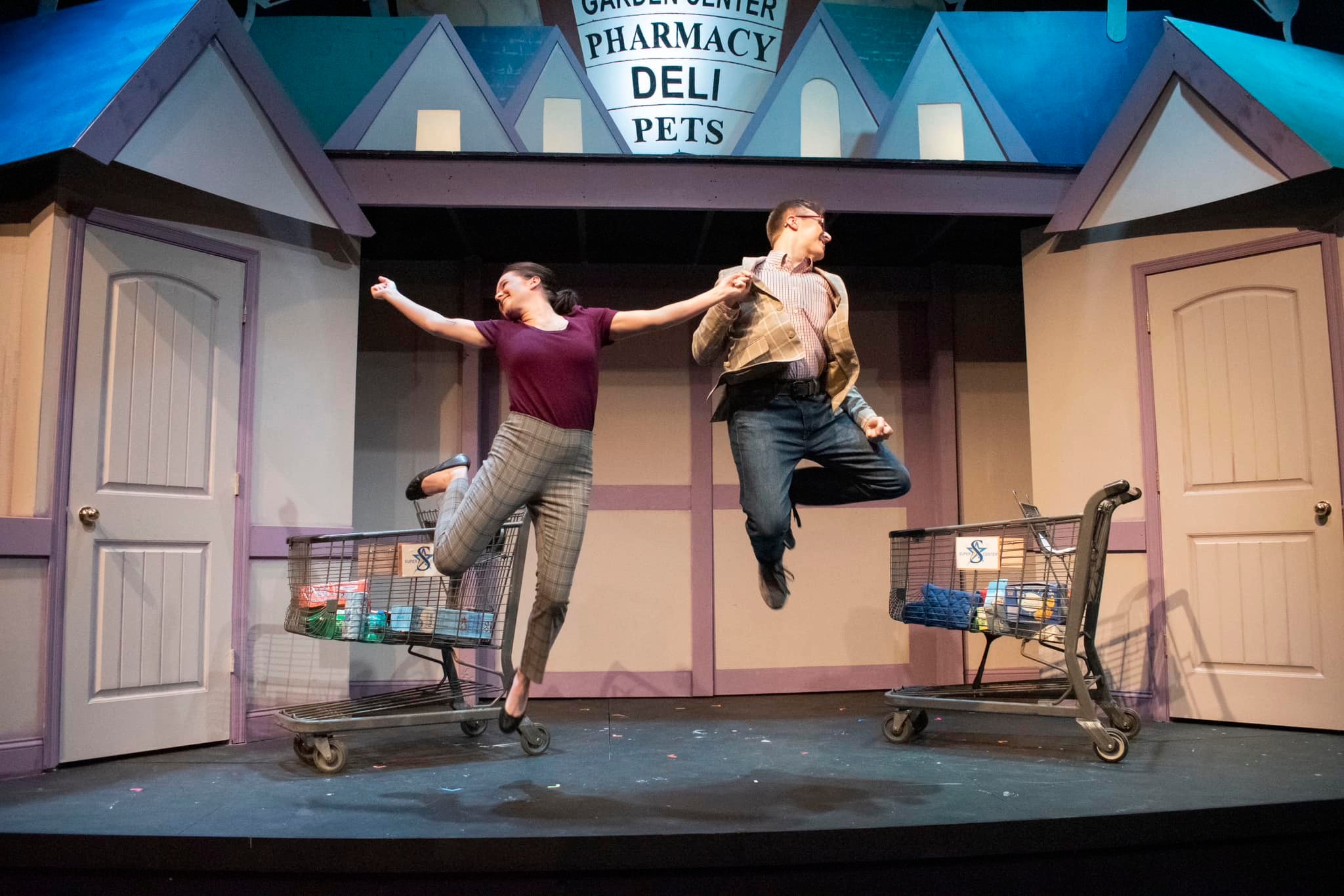 Two actors leaping into the air on a stage set to look like a grocery store, with two shopping carts and two doors on the left and right sides of the stage.