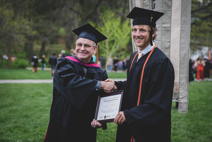 Doane professor and graduate in robe and graduation baps shaking hands while holding the grad's diploma.
