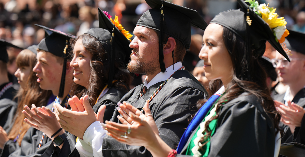 A row of graduates in black caps and gowns are standing and clapping, with rows of students and family members visible behind them.