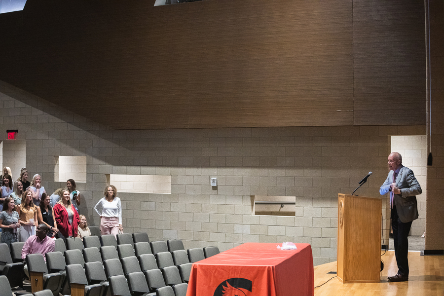 Dr. Roger Hughes reaches into the inside pocket of his grey suit jacket for his cell phone, to take a photo of Doane alumni who are employees of Crete Public Schools standing in the Crete High School auditorium.