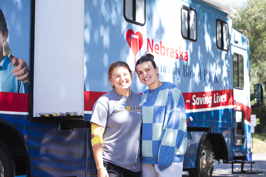 Two students, one with a yellow bandage around her elbow after donating blood, stand in front of the Nebraska Community Blood Bank's Bloodmobile outside of Perry Campus Center in Crete.