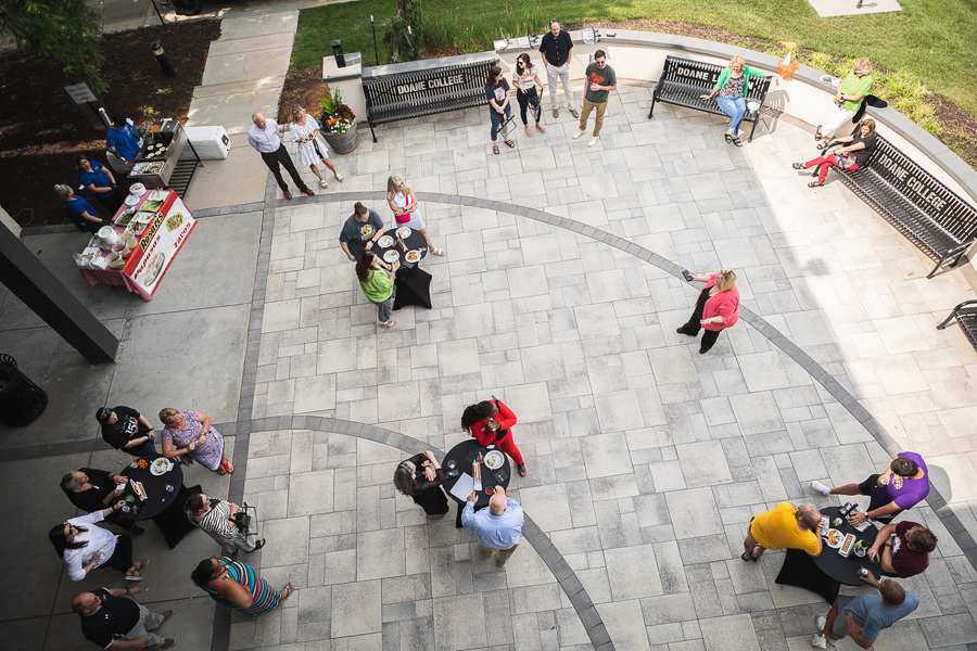 Anne Ziola speaks with Doane employees crowded around round cocktail tables on the patio of the Fred Brown Center. The image is looking over the patio from the third floor of FBC. 