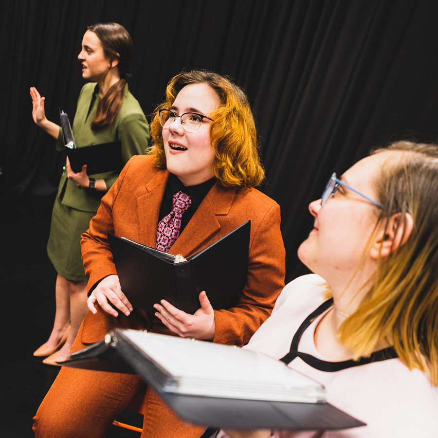 Student Olivia Vore sits on an orange stool in an orange pant suit, with a pink tie, holding a small black binder used for interpretive speech events. To their left stands Maddy Ramey in a green skirt suit. On their right, Ali Moulton stands in a pink suit and looks back at Vore. The three students are in front of a dark curtain on a black floor.