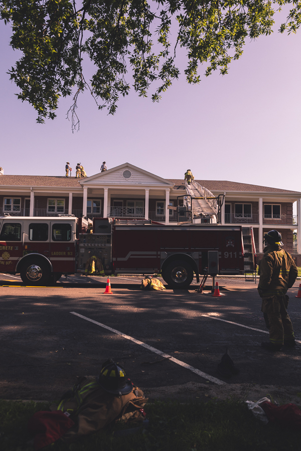 A firefighter watches from the ground as others practice cutting ventilation holes in the roof of Burrage Hall. Jackets and helmets are piled on the ground as other firefighters take a break from the June heat. 