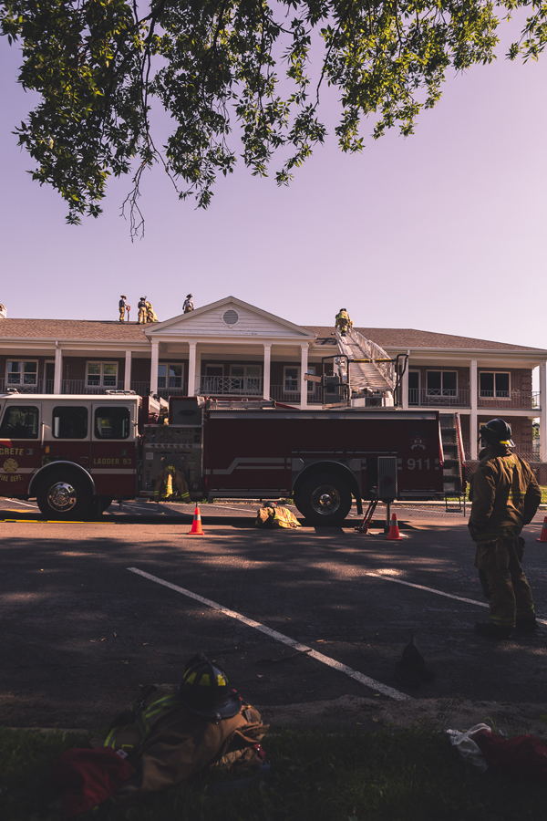 A firefighter watches from the ground as others practice cutting ventilation holes in the roof of Burrage Hall. Jackets and helmets are piled on the ground as other firefighters take a break from the June heat.