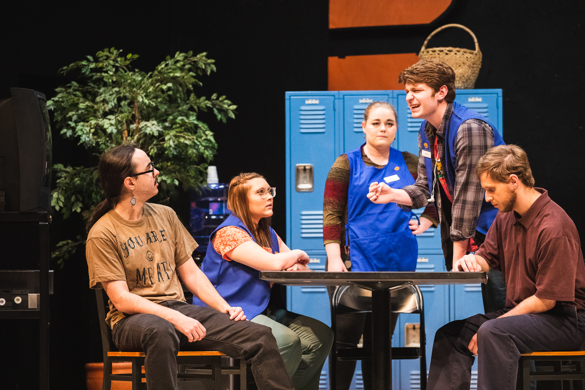 Five actors gather around a table, one standing over the table, clearly distraught. Behind them is a set made to look like the back room of a Hobby Lobby, with a tv on a stand, a sad plastic plant, bright blue employee lockers and a crafty-looking wicker basket. 