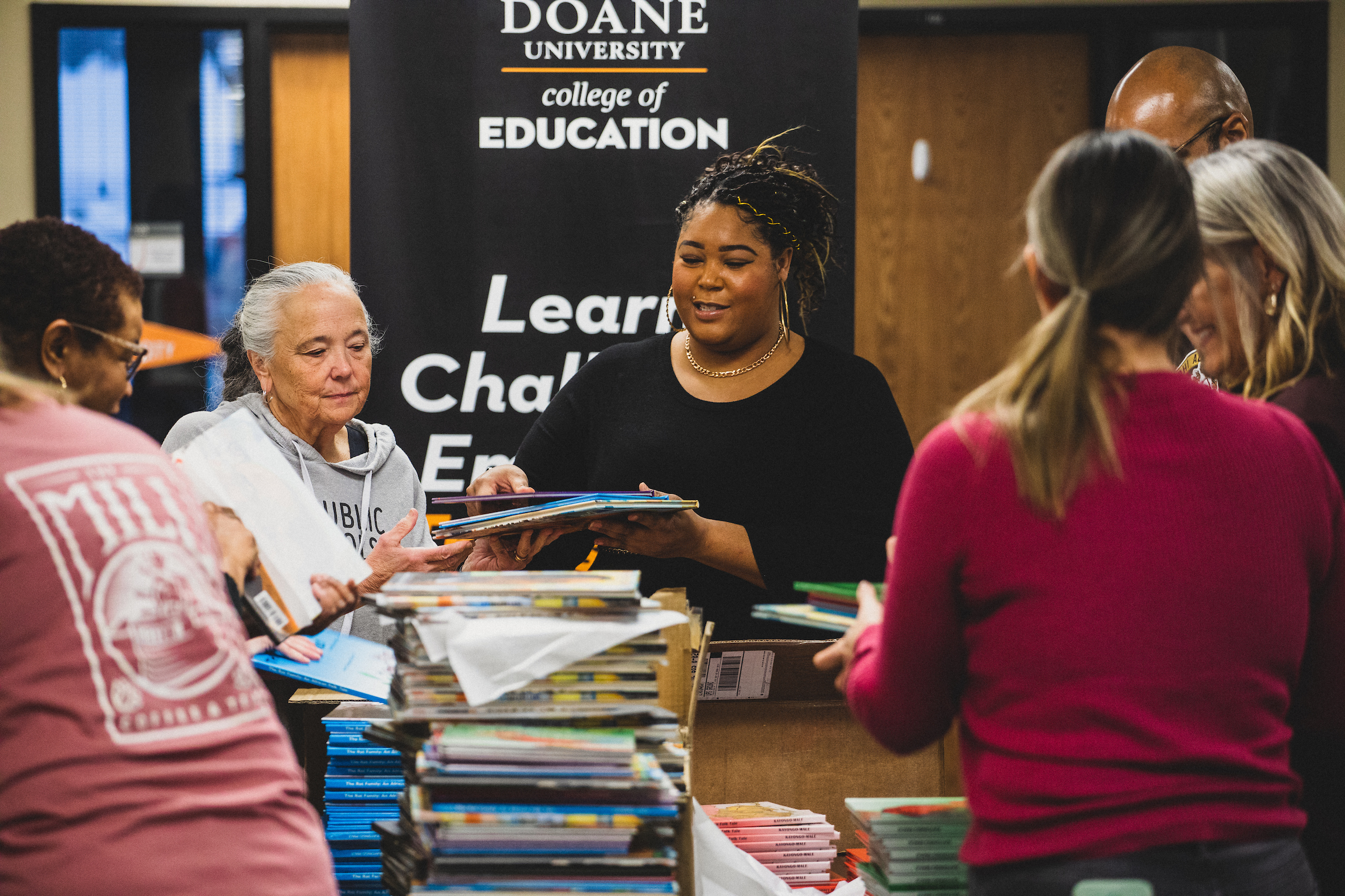 A group of people stand around a table gathering children's books into neat packs of five for donation. At the center stands Gabby Porter-Loving in a black dress, with a gold necklace and hoop earrings, and gold threads woven into her braids. She passes a stack of books to Dr. Bess Scott, who wears a grey sweatshirt that reads "I Love Public Schools." 