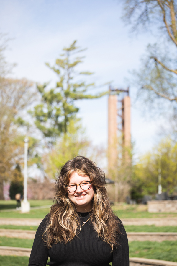 A portrait of Abrianna Miller from the grassy steps of the Cassel Theatre amphitheater. In the background is Merrill Tower, a thin brick tower with an open staircase winding to a bell at the top. Miller wears a black mockneck shirt, two gold necklaces and round tortoiseshell glasses. Her curly brown hair is parted in the middle and she's smiling.
