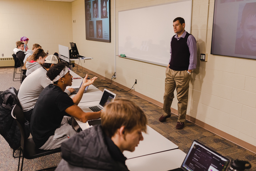 Dr. Timothy Hill, professor of political science, stands in front of a white board and two screens showing deepfake images. Students sit in a row at tables in front of him, with one student speaking and gesturing in discussion. 
