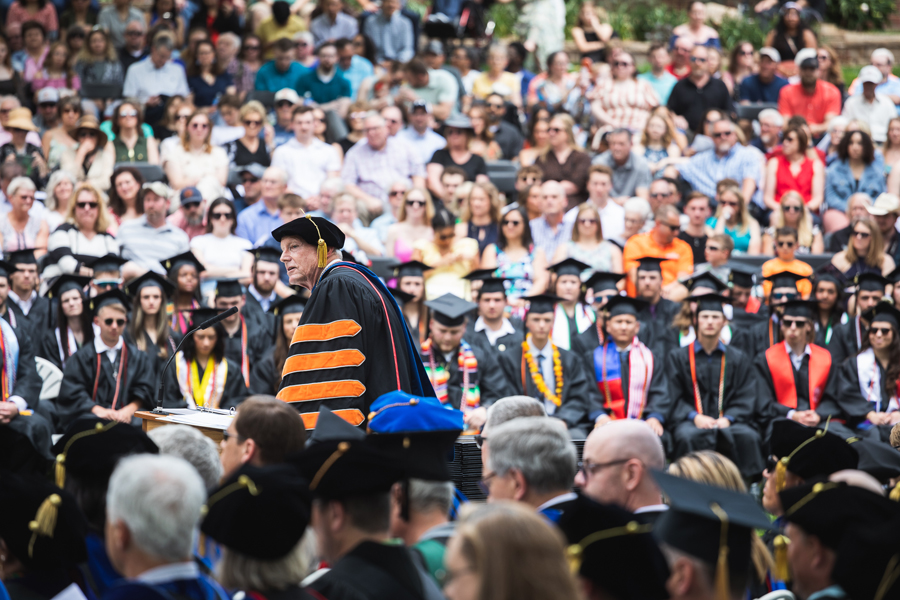 Doane President Dr. Roger Hughes speaks at a podium to a crowd of undergraduates waiting to receive their diplomas. In the foreground, members of Doane's faculty and staff are seated in full academic regalia, blurred, as are all the students and their families.