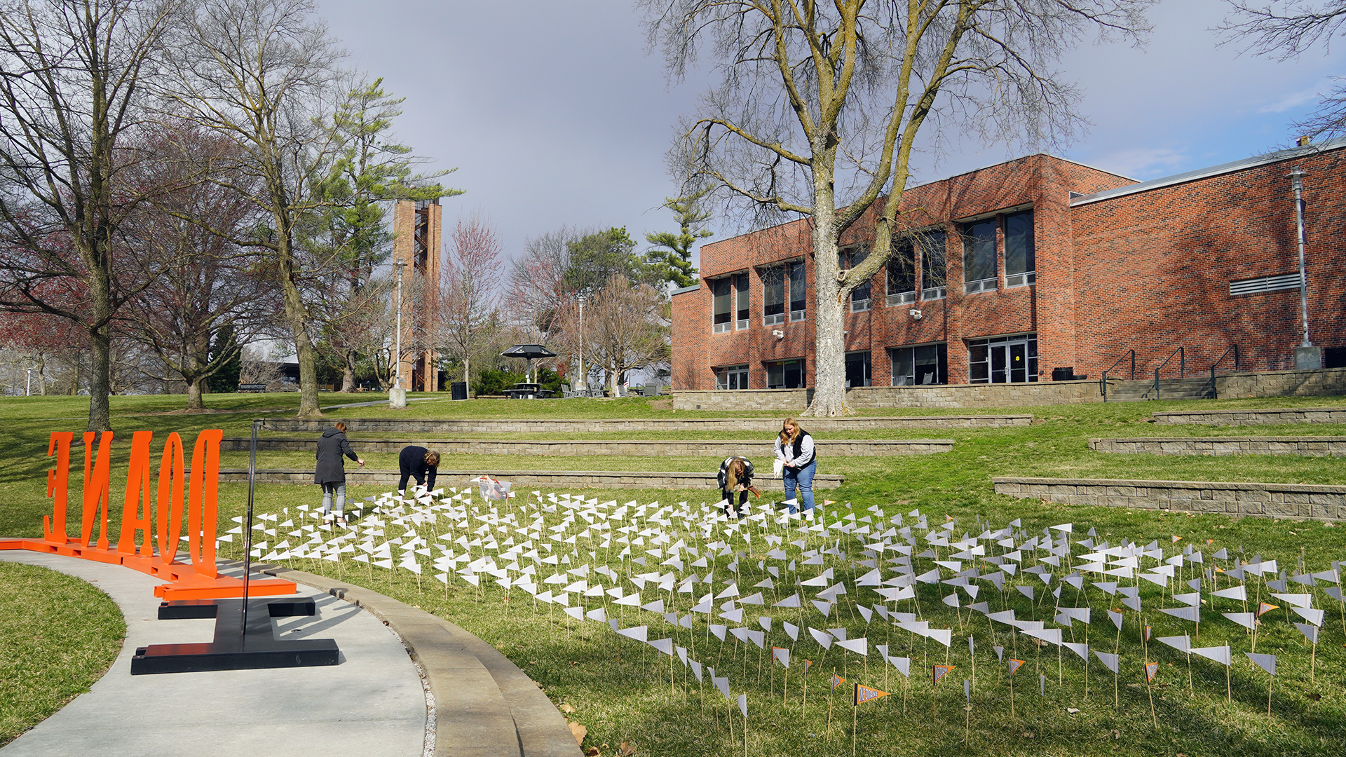 A group of staff members from the Advancement and Alumni Office stand inside Cassel Outdoor Theatre, placing one pennant per $200 dollars raised. Nearly 700 pennants can be seen in a tight area at the front of the amphitheater. The brick building known as Campus Center and the brick Merrill Tower can be seen in the background, as can a dark blue sky.