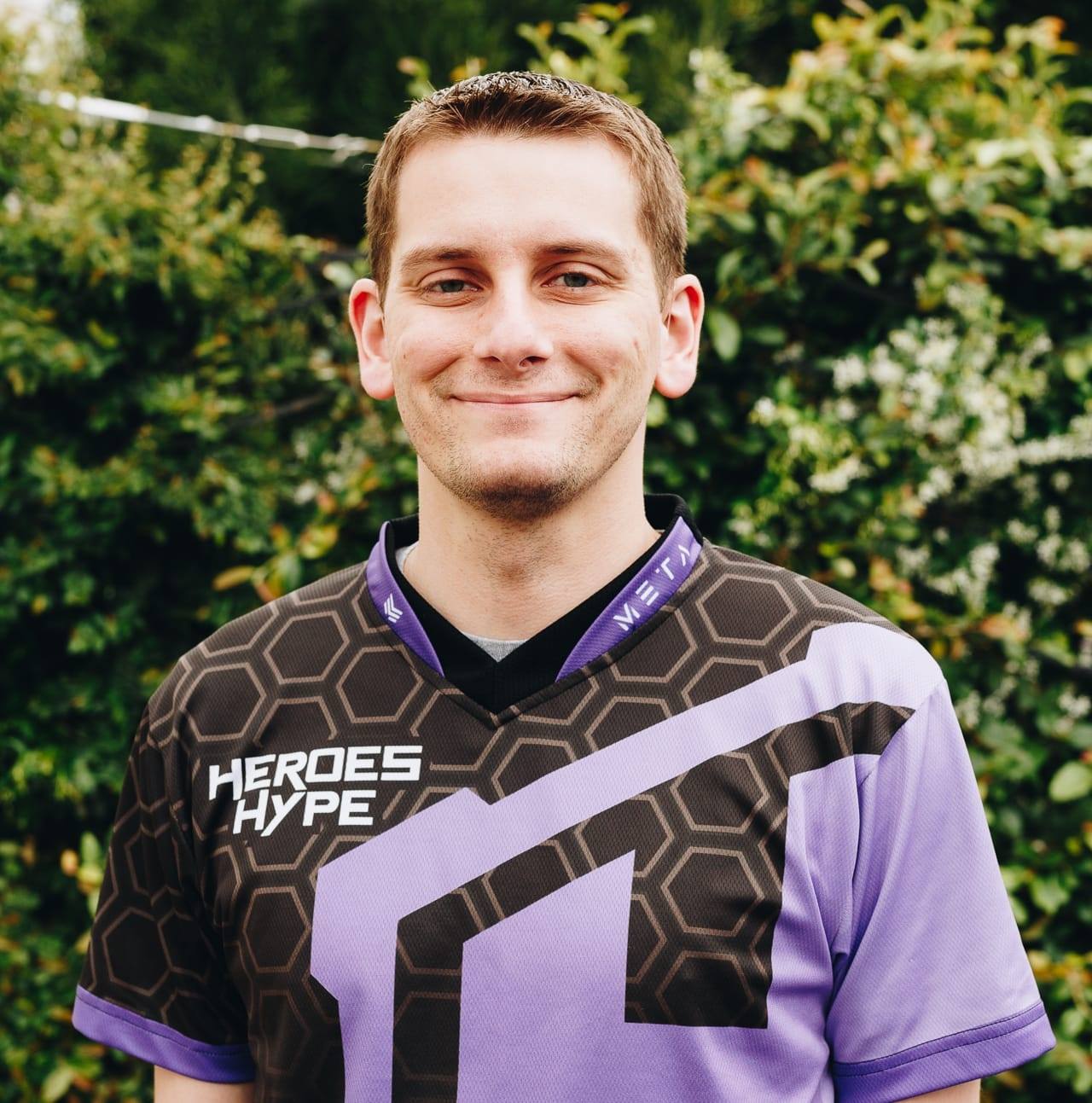 Image of Aaron Blackman, director of esports and head coach. He wears a black and purple jersey that reads "Heroes Hype," and stands in front of out-of-focus shrubs and bushes. His light brown hair is cut short and gelled up at the front. 