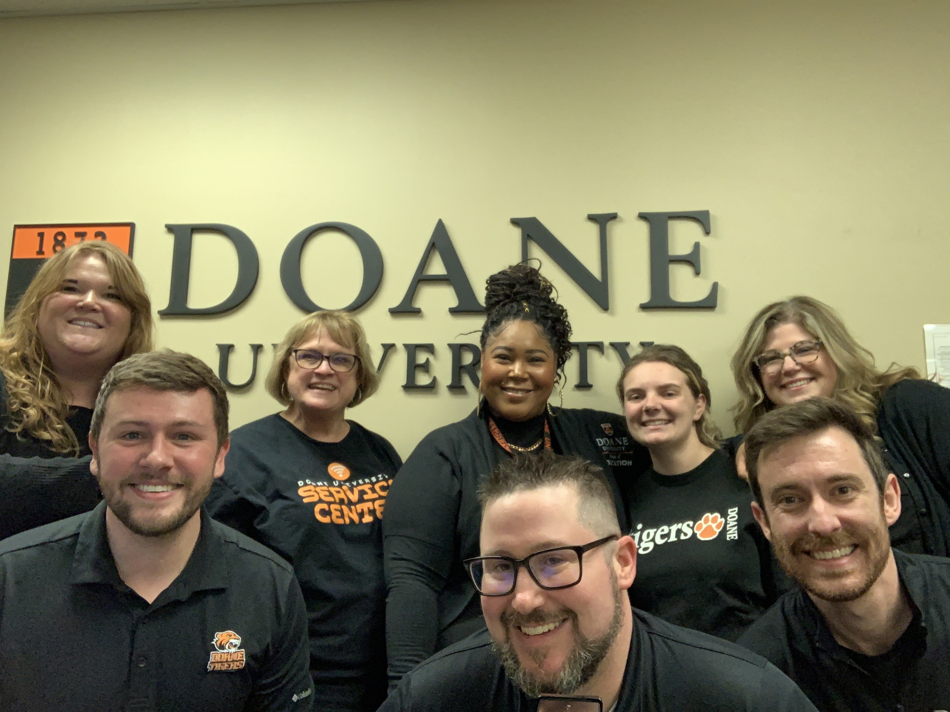 A group of employees from the College of Education, Recruitment, IT and the Office of Strategic Marketing stand in front of the Doane University sign inside the Administration building on Doane's Lincoln campus. 