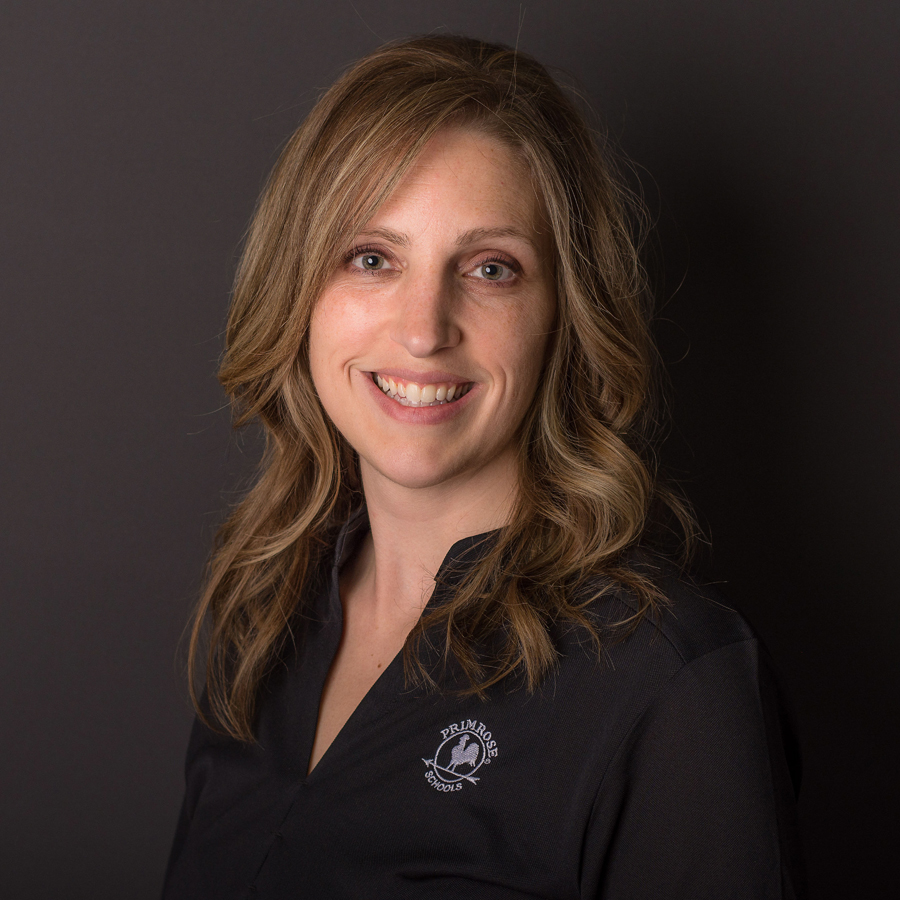 An image of Dr. Betsy Tonniges, who has long, curled brown hair and wears a black jacket with a white embroidered logo in the shape of a rooster on an arrow — the logo for Primrose Schools. The photo has a dark grey background.