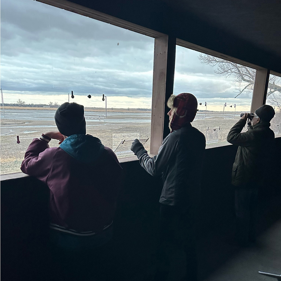 Jeremy Caldwell opens a small door cut into a plexiglass window in a crane-viewing blind on the banks of the Platte River. To his left is a Doane student, and a bird enthusiast uses binoculars on his right. The day is cloudy and a dusting of snow is collected at the edge of the grass along the riverbank.