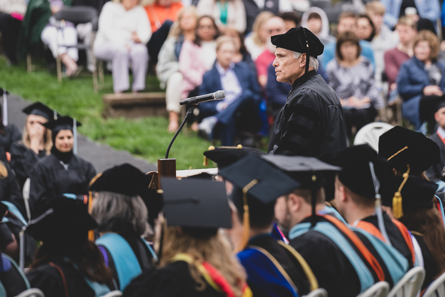 Dr. Steve Joel stands on a podium above the heads of Doane faculty and staff in full academic regalia in Cassel Theatre for the spring Commencement ceremony. Beyond him are the blurred faces of graduates and their families. 