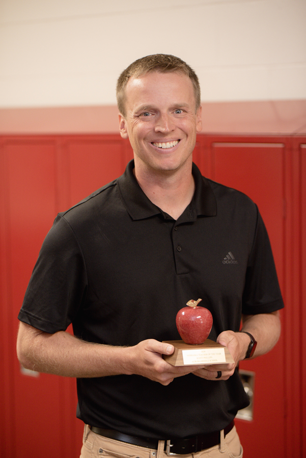 Scott Phillips holds the award declaring him the 2024 Nebraska Teacher of the Year. He wears a black polo tshirt and holds the award, a red granite apple on a wooden base, just in front of his stomach. Behind him are the red lockers of Aurora Middle School. 