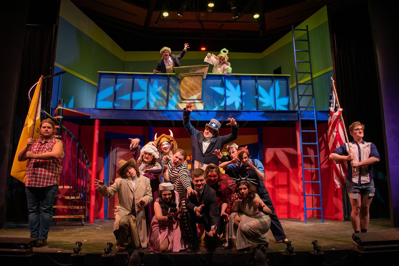 An image from the production of "Reefer Madness" showing actors on a two-level set with brightly colored lighting shaped to look like marijuana leaves. Two actors, one in a fur cap with a tail and one in an angel costume, stand on the top level of the set. On the base level, two actors stand on the far left and right with flags, and a group of 11 actors pose together in the middle. 