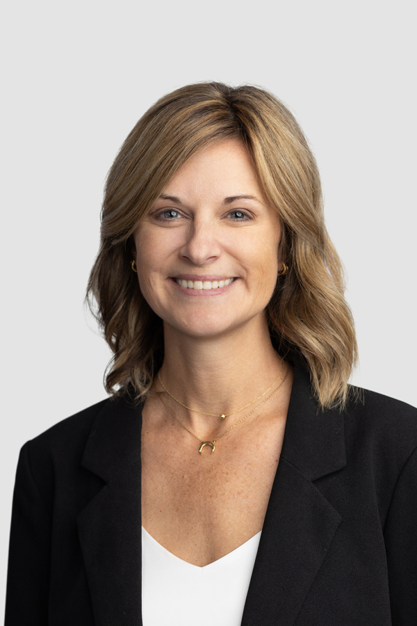 Image of Darcy Dawson, director of Doane's Counseling Center. Dawson has shoulder-length dark blonde hair with a smile, and wears a black suit jacket over a white v-neck shirt with two gold necklaces. The background is white. 