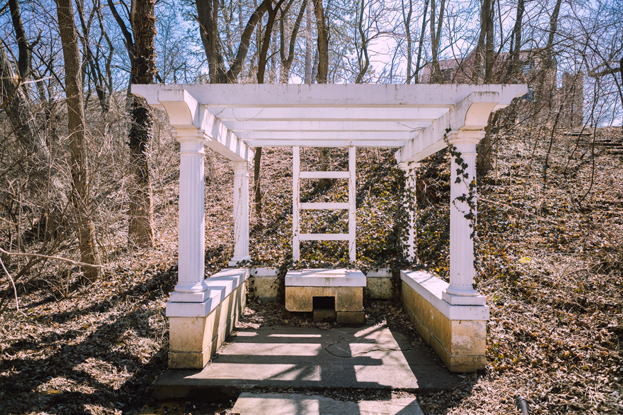 An image of Dean Pergola, a Greek-inspired pergola with white fluted columns situated around a now-capped spring. The image is from early spring, so the trees are bare and Frees Hall can be seen in the background at the top of a hill.