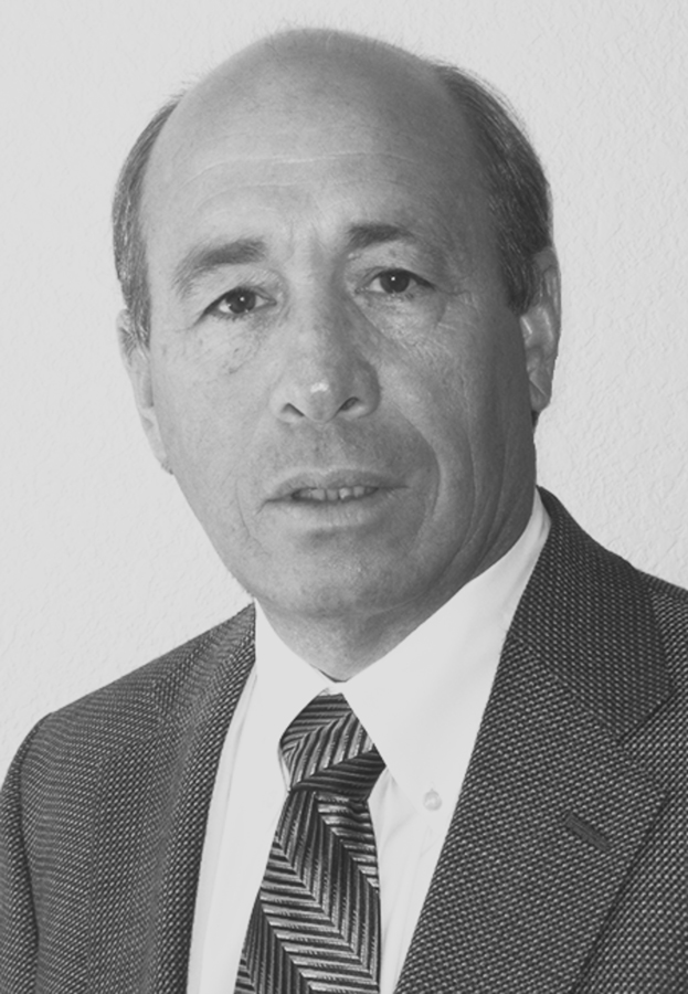 A black and white headshot of Dick Shoemaker, who wears a white button-up shirt with a zig-zag patterned tie, and a woven suit jacket. 