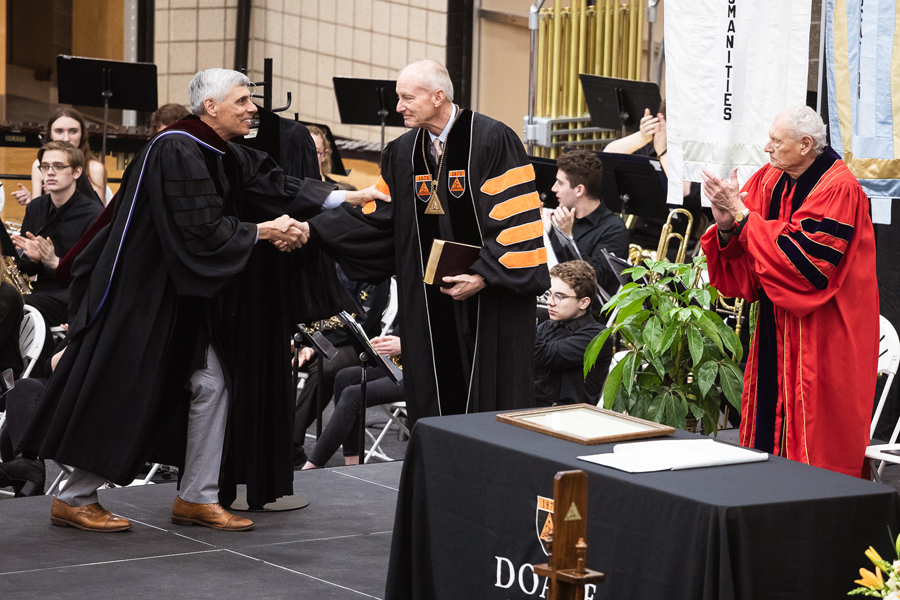 Rev. Dr. Jim Keck reaches to shake the hand of Dr. Roger Hughes on a stage during the inauguration of Hughes as Doane University's 13th President.