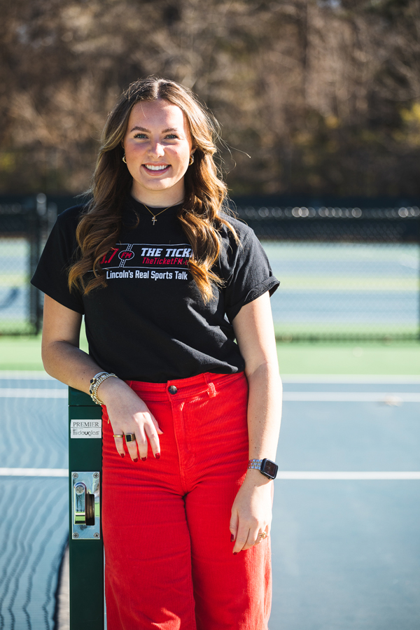 Image of Grace Schroller, leaning against a green pole for the tennis nets on Doane's blue tennis courts. Schroller wears a black shirt that reads "93.7 The Ticket" and red corduroy pants.