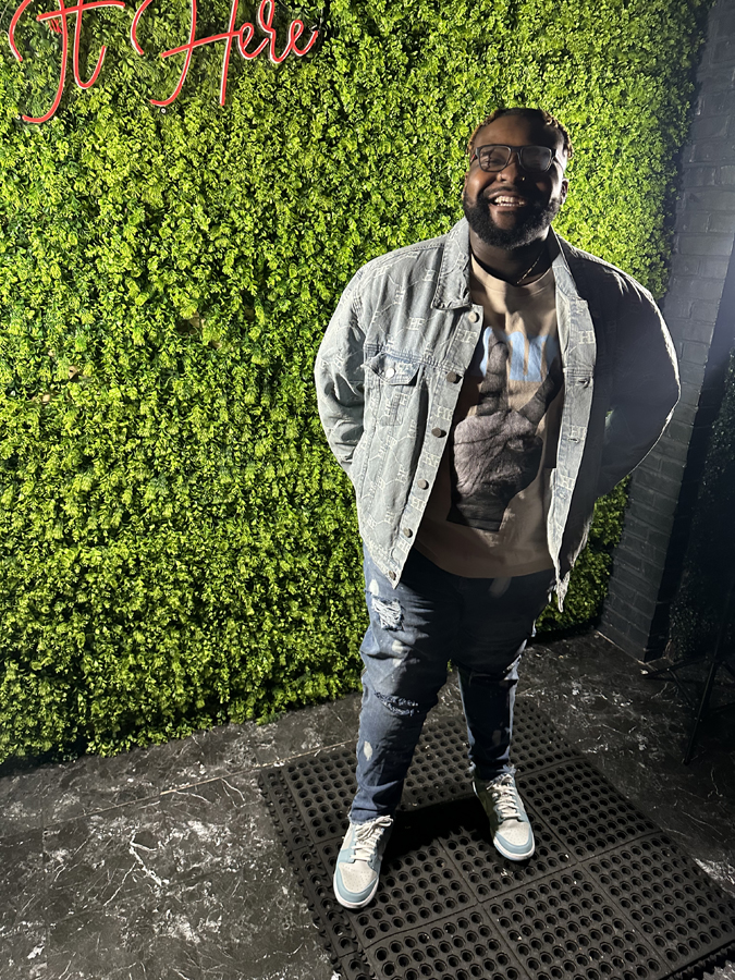Eric Jones stands in front of a wall with faux greenery and black-painted brink, wearing Nike tennis shoes, jeans and a denim jacket. 