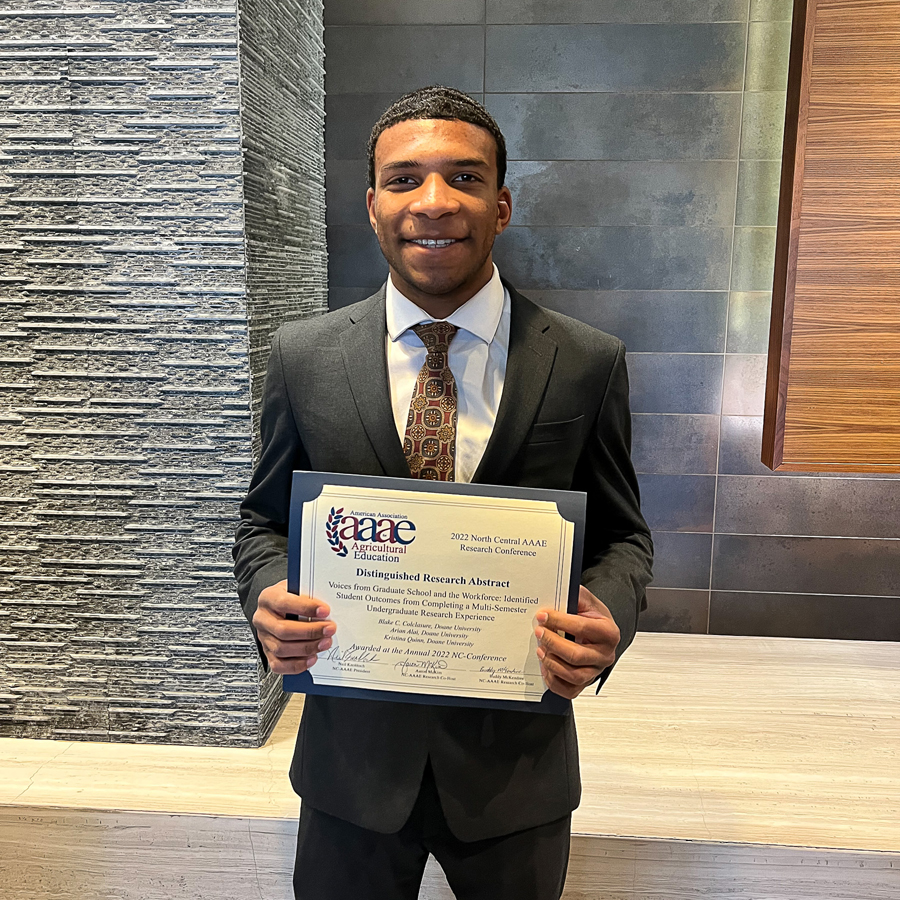 Arian Alai ’23 holds the award recognizing the abstract for "Voices from Graduate School & the Workforce" as one of the distinguished papers at an October conference. Alai is wearing a grey suit and white shirt, with a colorful tie. 