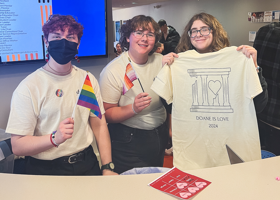 Three students stand behind a counter, one holding a rainbow Pride flag, one holding a lesbian Pride flag and one holding up a cream-colored shirt. Printed on the shirt is a series of four broken and whole pillars surrounding a heart, under which reads "Doane is Love 2024."