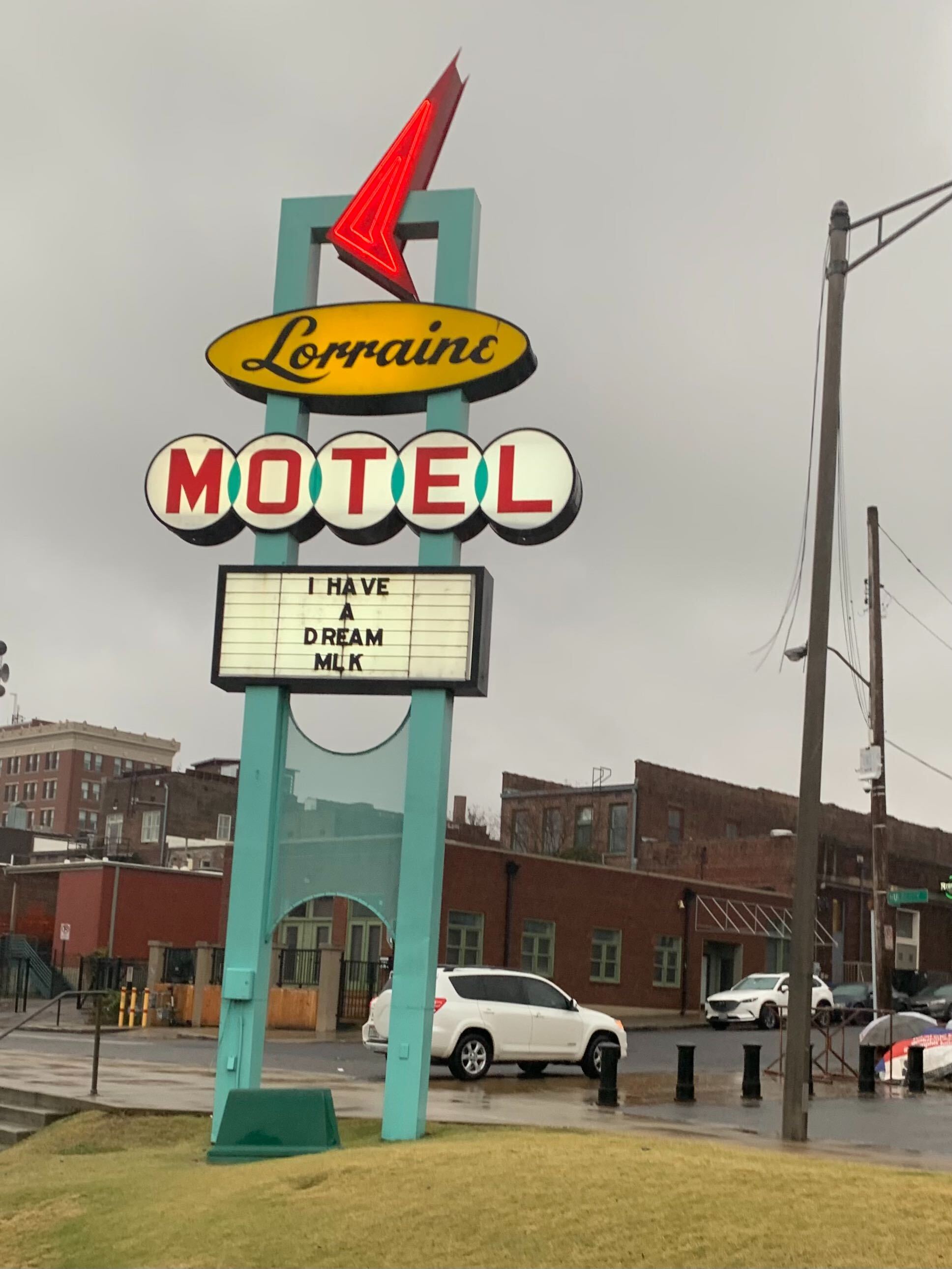 A photo on a rainy day showing the sign for the Lorraine Motel, which has a rectangular turquoise frame topped with a stylized red neon arrow. The text for Lorraine is in a cursive script inside a yellow field underneath the arrow, with "motel" underneath still in red block letters inside white circles. A board below the letters reads "I have a dream — MLK." 