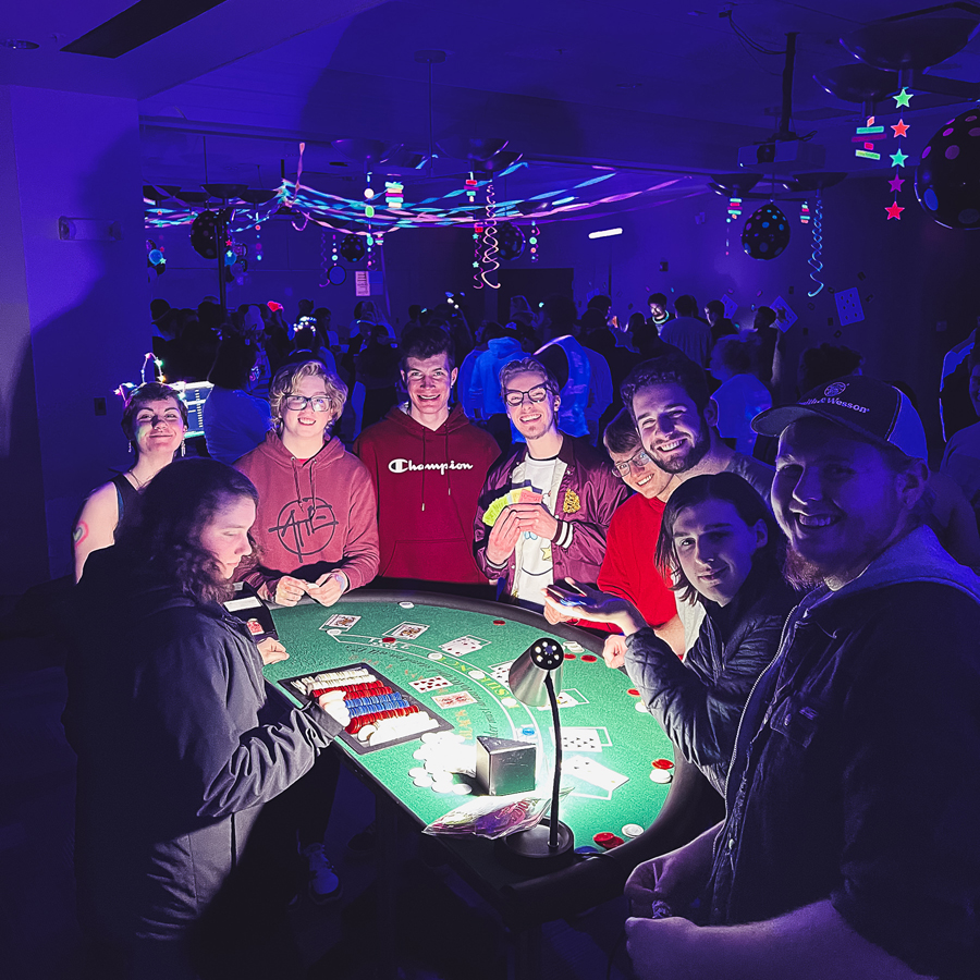 Doane students gather around a blackjack table lit by a small lamp. The rest of the room is lit by purple-hued blacklights and glows with neon streamers and decorations.