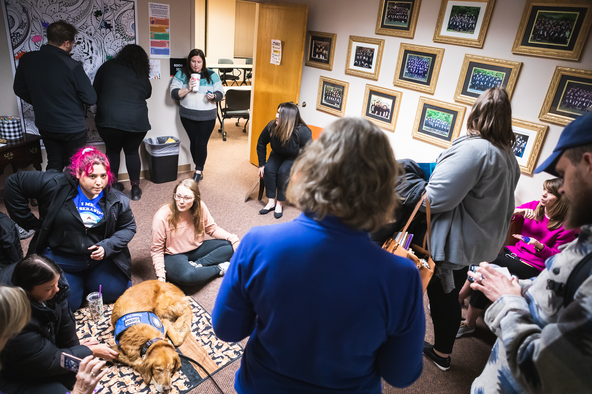 A group of people engage in activities around a small room, talking, drawing and petting a dog laying on the carpet. 