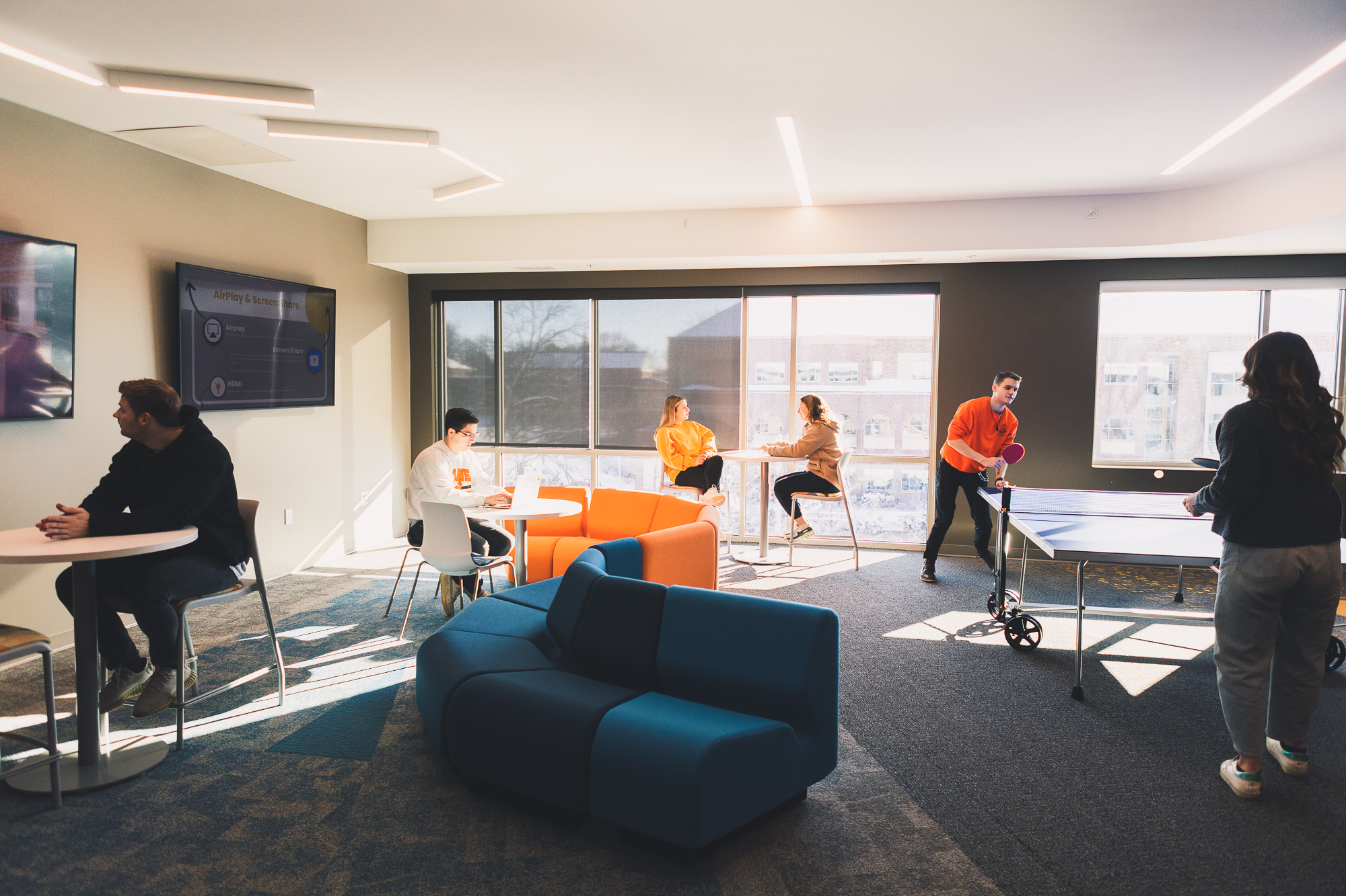 Students use a lounge in New Hall, which is well-lit by a bank of windows. Two students play ping-pong to the right, while two other students sit and talk at a high-top table near the windows. A third student works on a laptop at a small, white, round table around which an orange couch curves. 