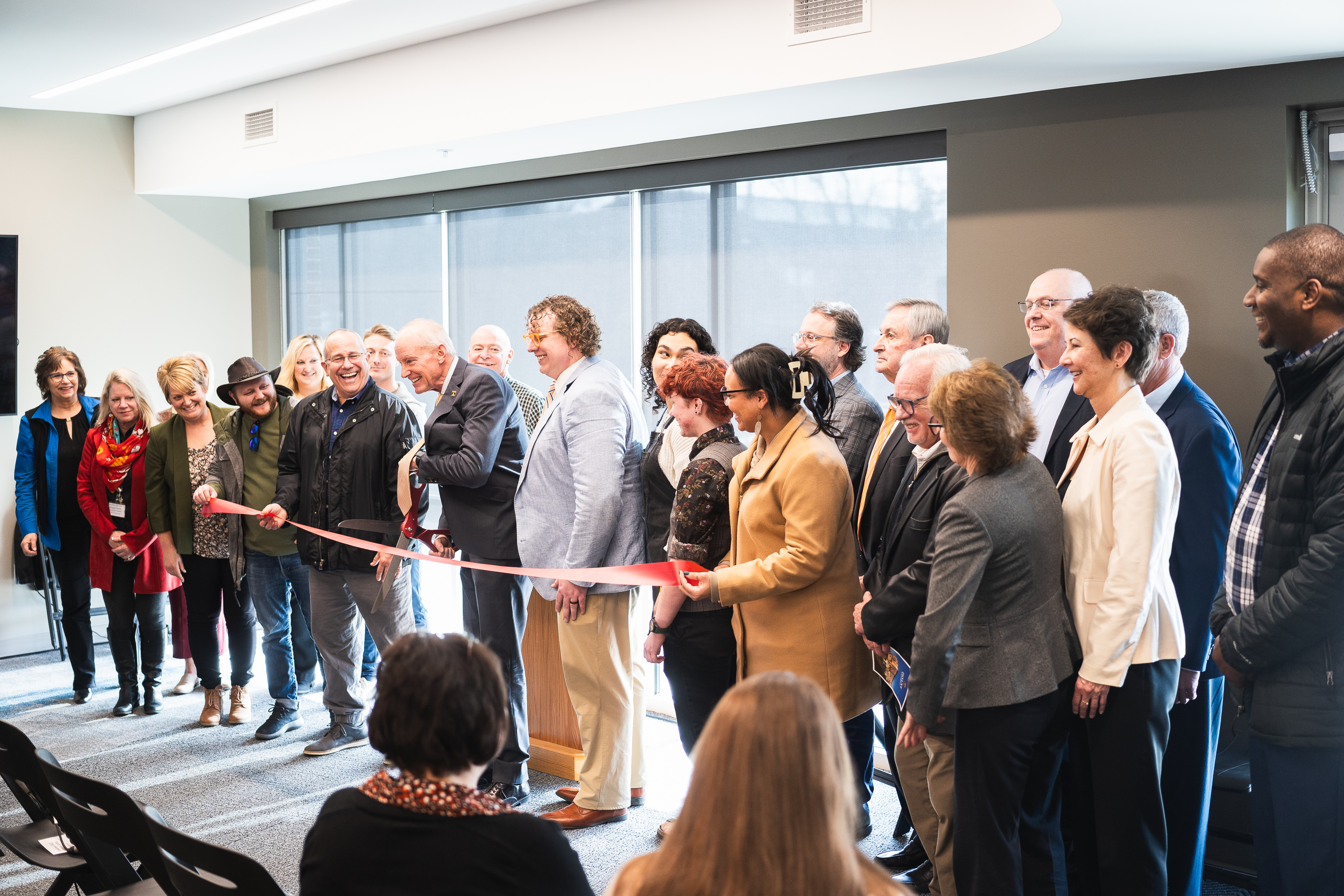 A group of people hold a red ribbon, with one in the middle holding a pair of large scissors to cut the ribbon. They stand indoors, in front of a wall of windows. 