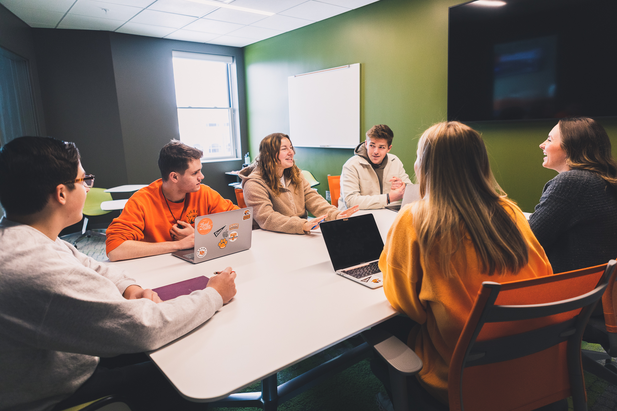 Six students sit around a white, rectangular table in a vibrant green-painted study room. Behind them are individual desks and chairs, and a window. 
