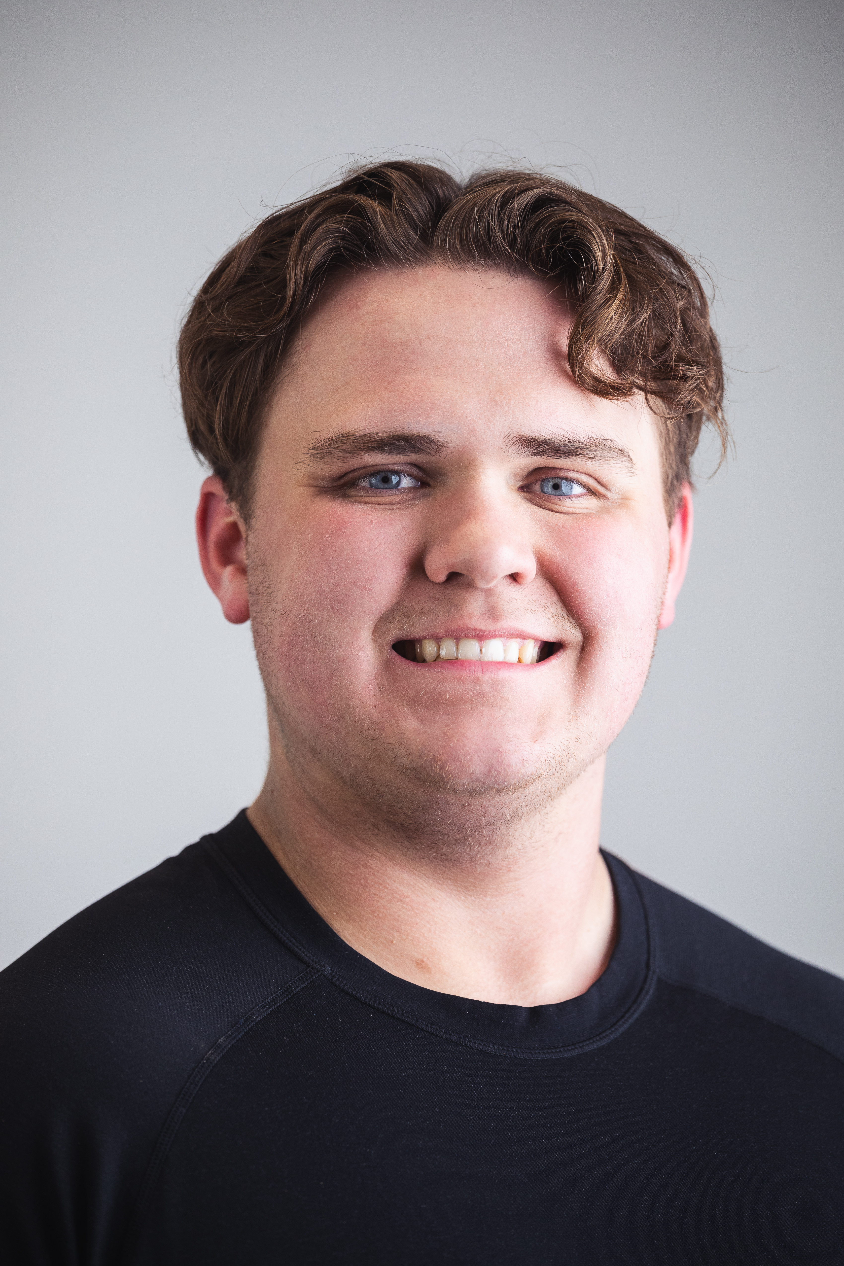 Johnathan Schmidt wears a black tshirt. He has short brown hair with a couple curls that drop over his forehead and blue eyes.