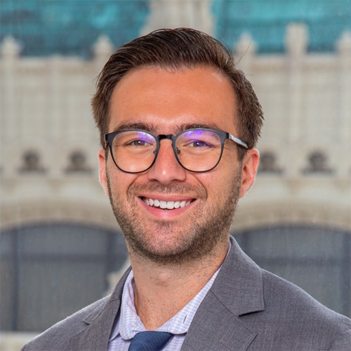 Image of Travis Handler wearing large, round, black-framed glasses, a blue and white houndstooth shirt and grey blazer. In the background is a gothic-style building with a green roof.