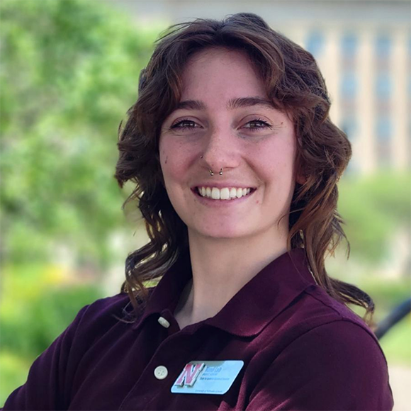 Image of Mandi Laib, wearing a dark red, short-sleeve polo with their University of Nebraska-Lincoln nametag near the collar. In the background is Love Library, on UNL's city campus.   