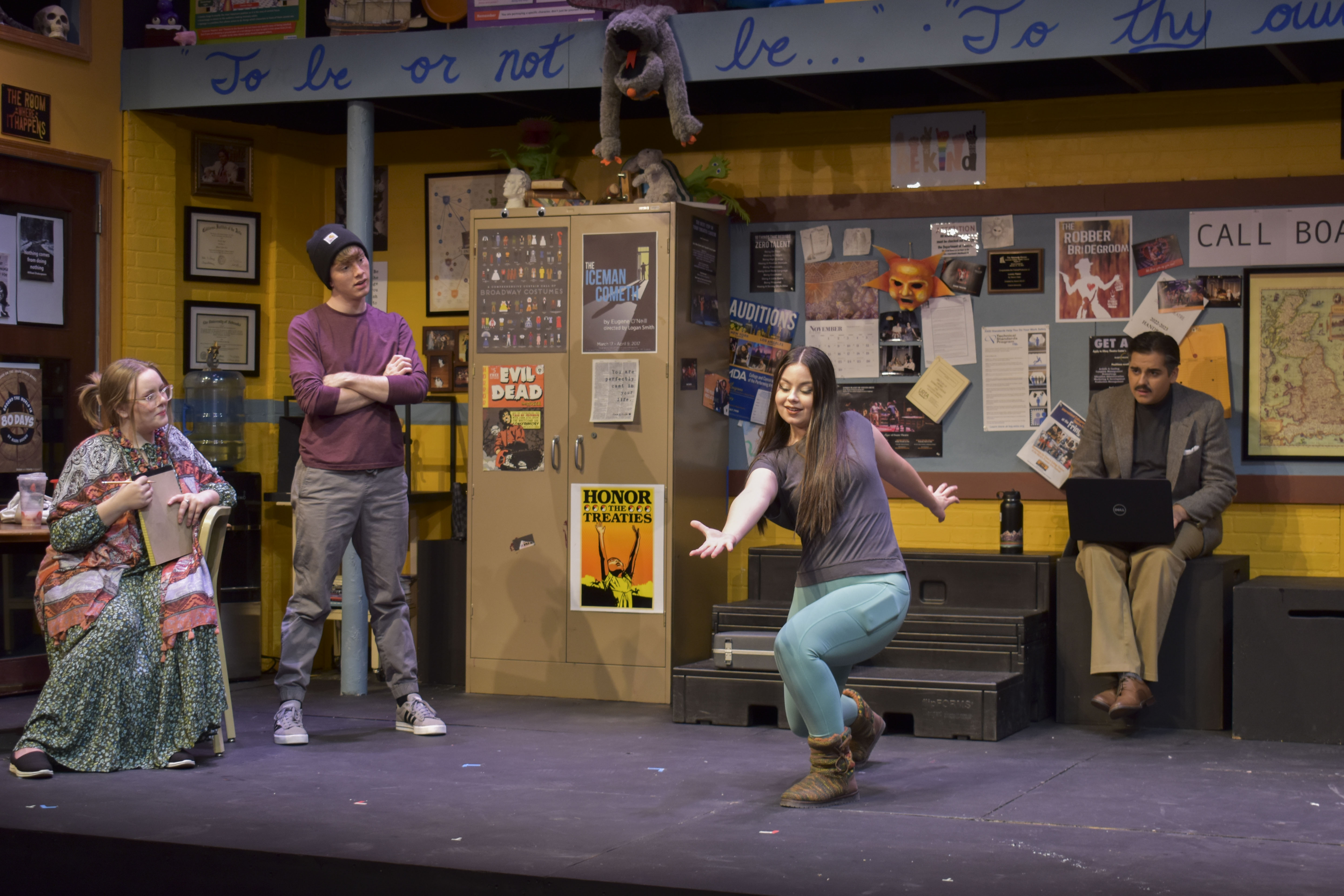 Four actors pose on a stage, with one miming a bowling motion. The stage has two levels, and a puppet dangles precariously from the second above the words "To Be or Not To Be." The rest of the set looks like a theatre classroom, with a desk and chairs, posters covering the walls and props littered around the edges.