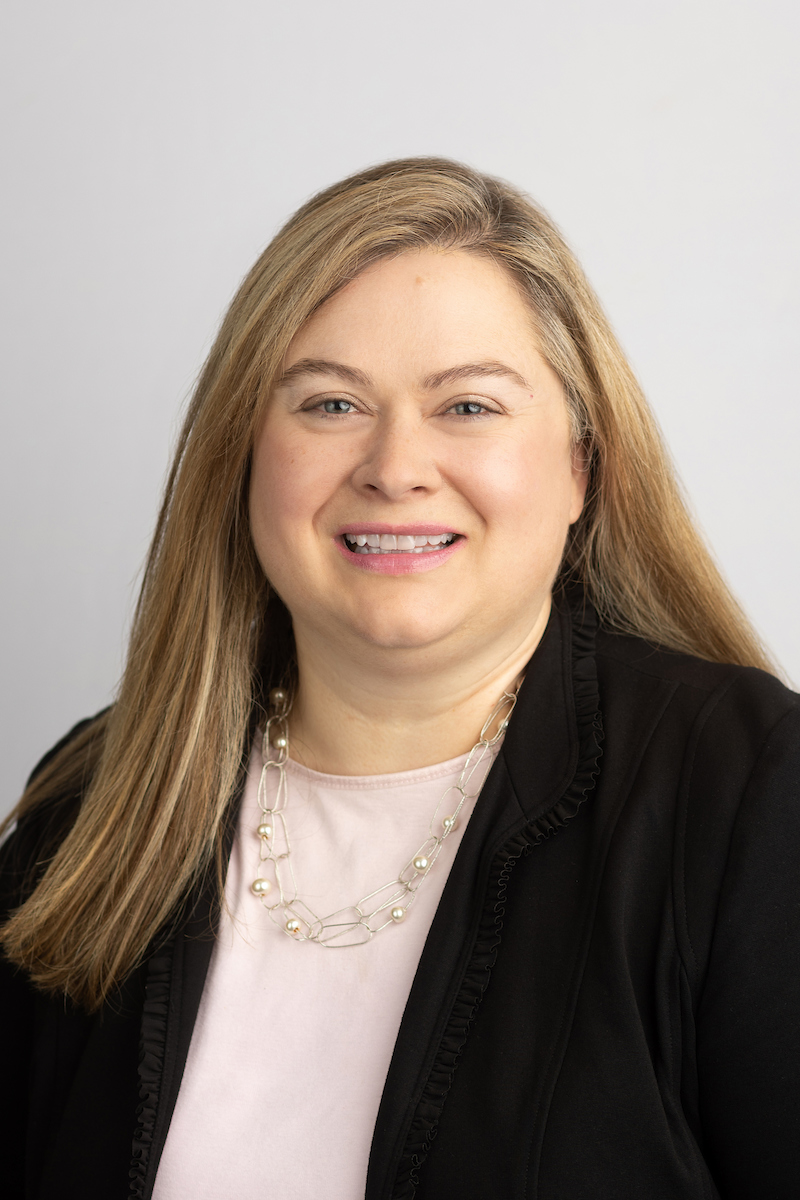 Portrait of Dr. Jen Bossard, who has long blonde hair and is wearing a black jacket over a pale pink top and necklace.