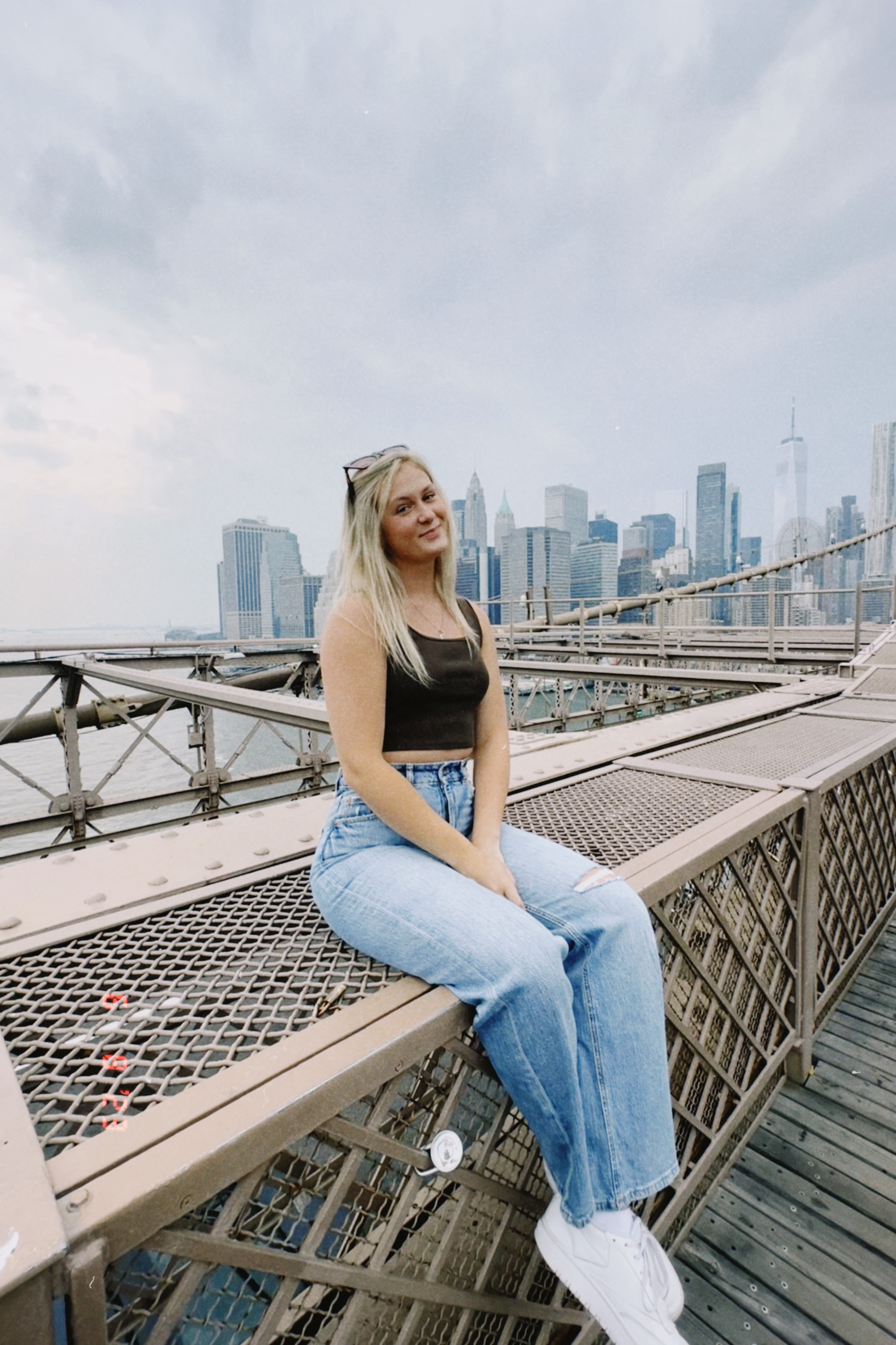 Image of Blair Souchek in a black cropped tank top and light-colored, high-waisted jeans, seated on the walking path of a bridge overlooking the New York city skyline. 