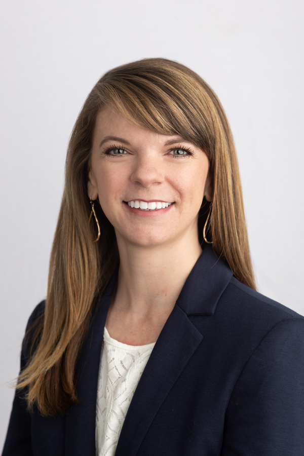 Image of Dr. Courtney East, who has long brown hair with highlights and a smile, wearing a navy suit jacket over a lace shirt. The background is white.