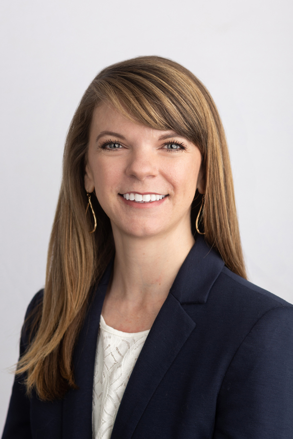 Image of Dr. Courtney East, who has long brown hair with highlights and a smile, wearing a navy suit jacket over a lace shirt. The background is white. 