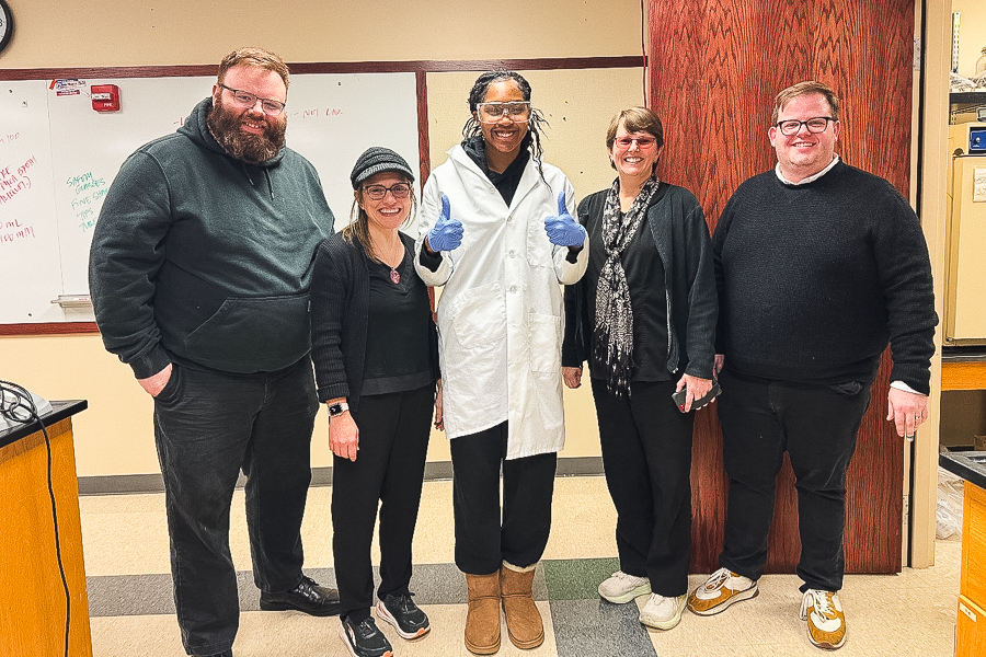 Five people stand in the doorway of a laboratory classroom, all wearing black. The student in the center has her hands raised in a double thumbs-up and wears a white lab coat. 