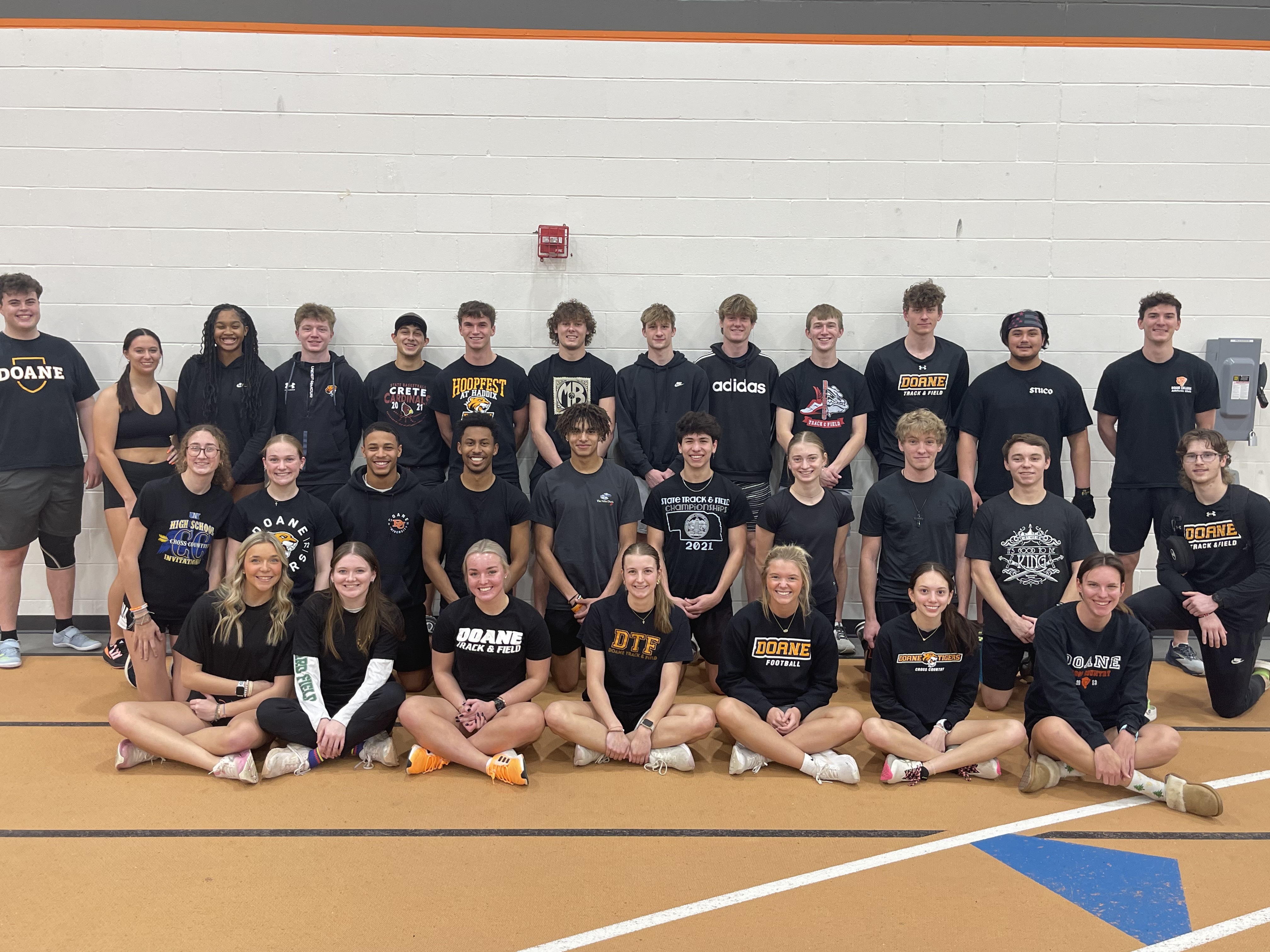 An image of 30 members of the Doane Track & Field team standing or seated in three rows on the track inside of Furhrer Fieldhouse. All 30 students are dressed in black pants and tops.