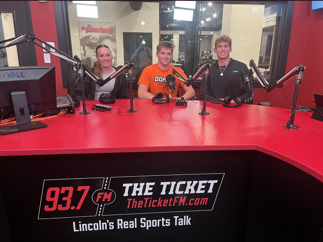 Grace Schroller sits behind the microphones in a recording booth at 93.7 The Ticket with fellow student-athletes Zach Fye and Guy Moran. The top of the desk and walls are painted Husker red, and the base of the desk is black. Behind the three students are black-framed windows and a glass door that lead to the rest of the studio.