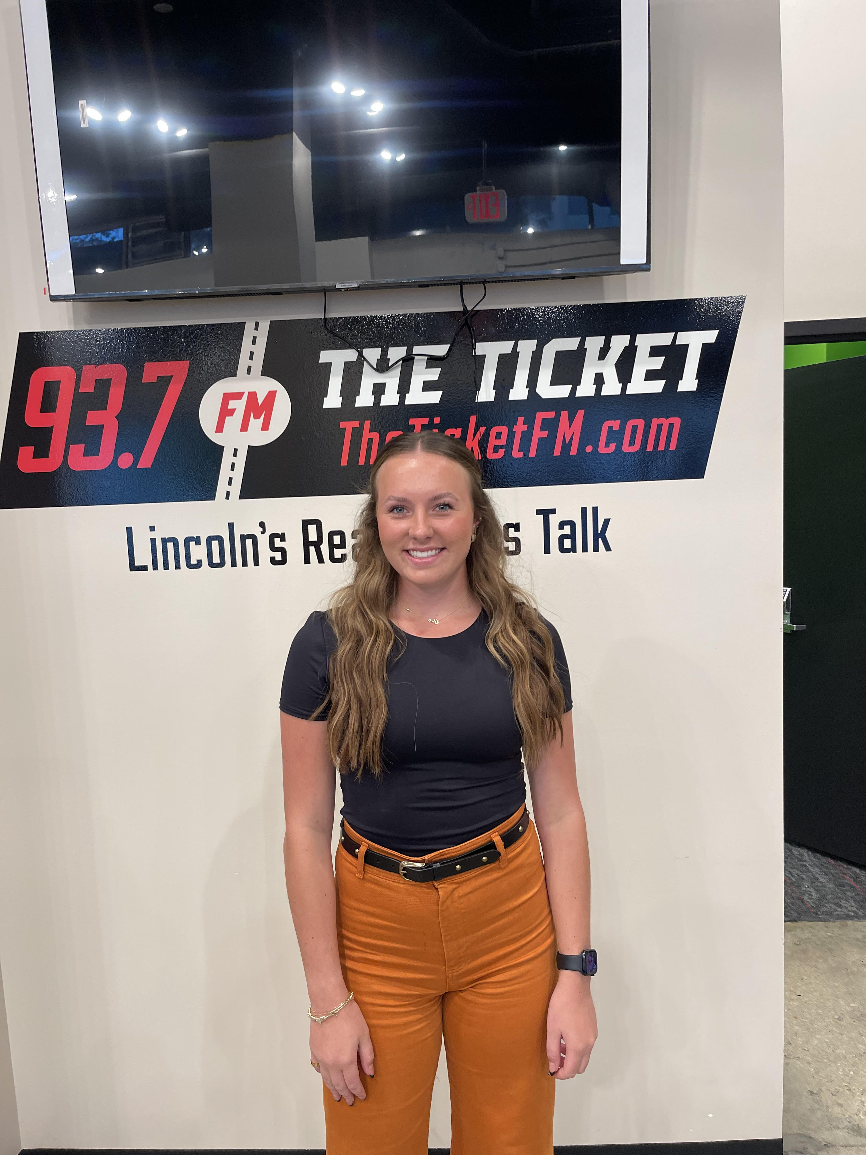 Grace Schroller, who has long, curled brown hair and wears a black tshirt and orange pants, stands in front of a sign for 93.7 The Ticket.